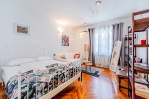 A hotel room with a bed, a bedside table, a lamp, a mirror, a shelf with books, and a window with curtains. - Cozy Studio in the Center of Zagreb (Zagreb)