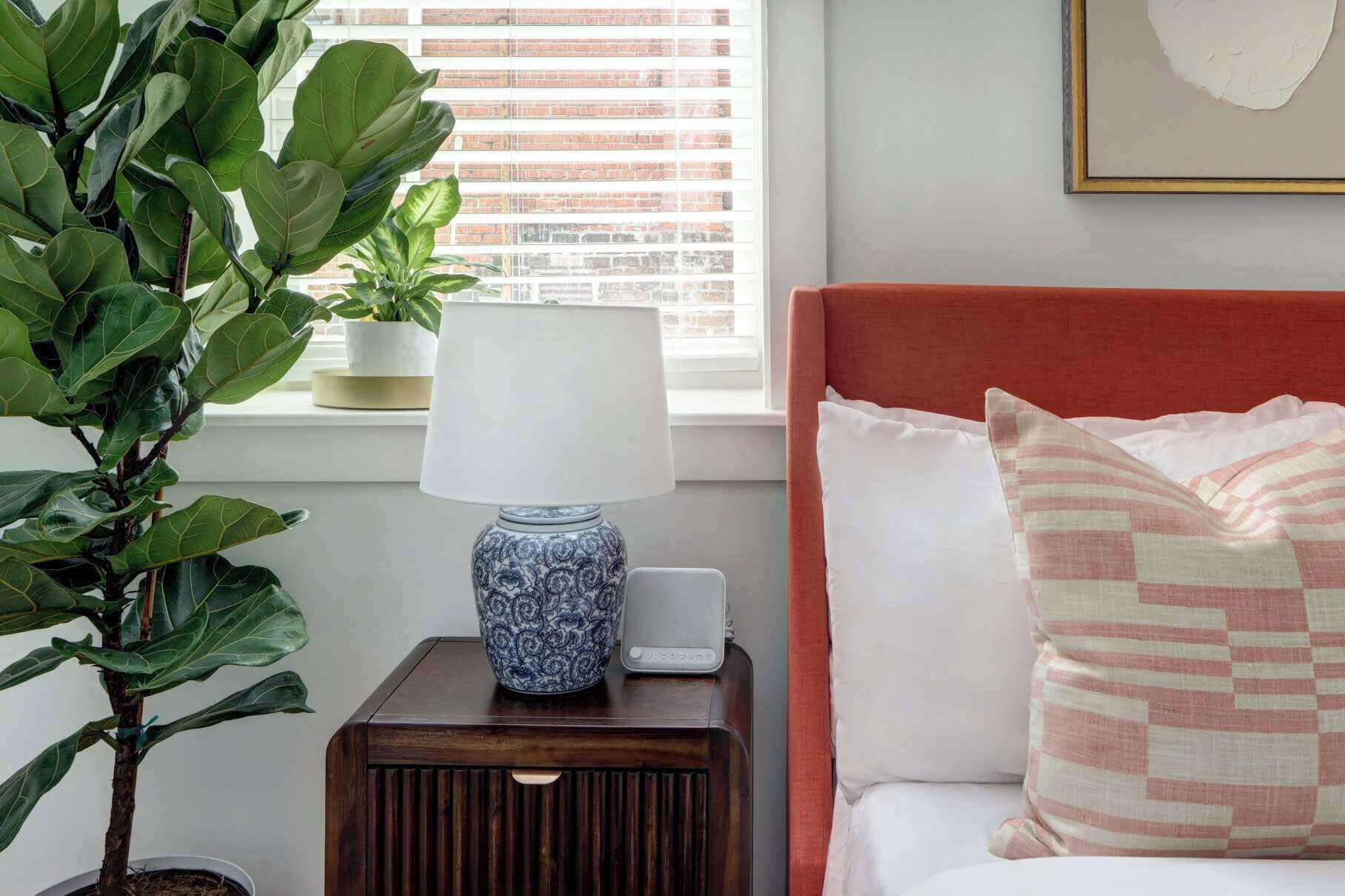 A bedroom with a bed, a bedside table with a lamp, a potted plant, and a window with blinds.