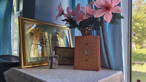 A table with a framed Quran, a decorative vase with pink flowers, and a small bottle. - Gabala Garden Hostel (Gabala)