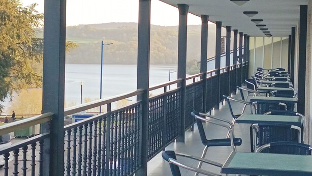 A covered outdoor seating area with tables and chairs, overlooking a body of water and hills. - Hotel Villajardin (Portomarin)