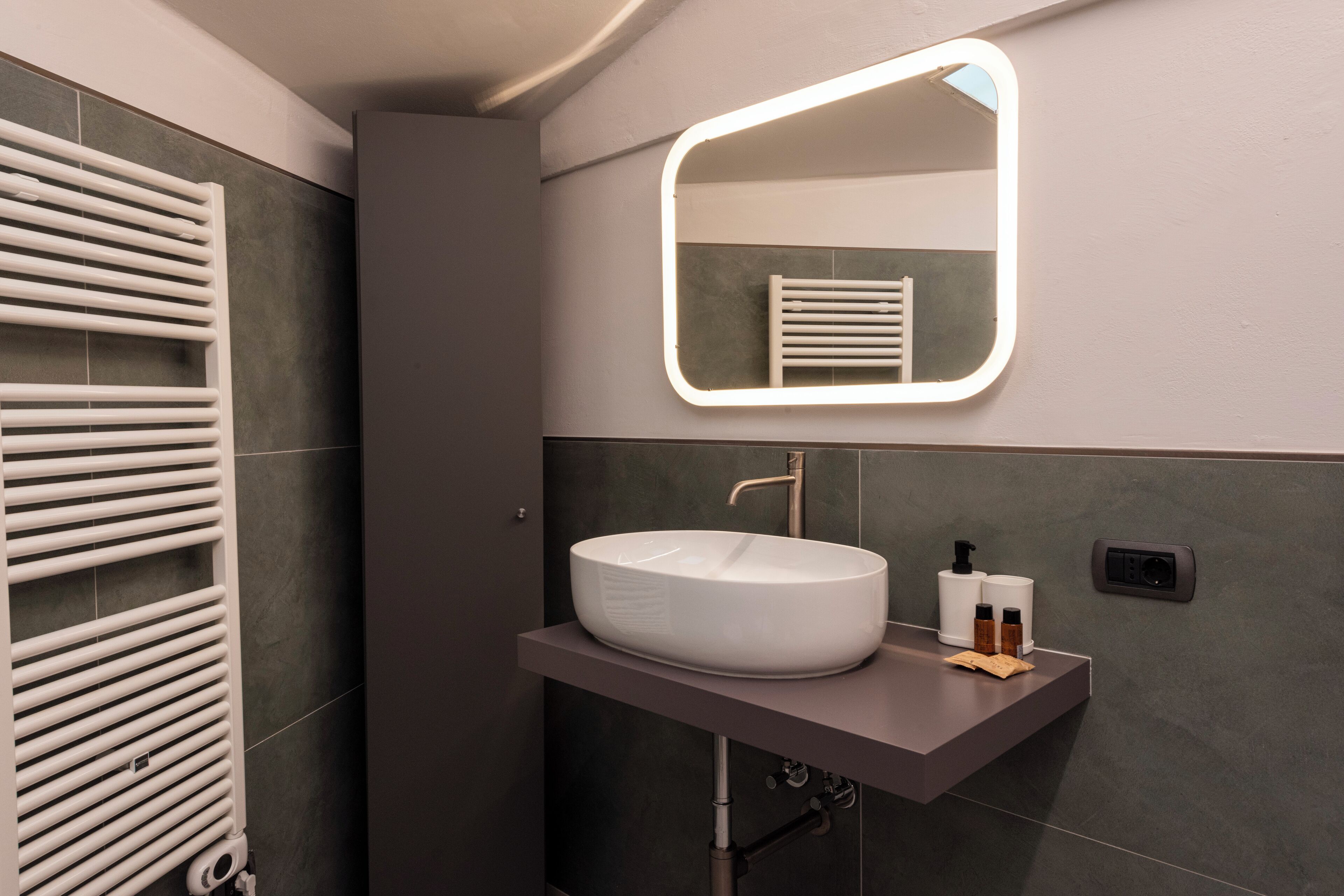 A modern bathroom with a white vessel sink, a towel rack, and a mirror with built-in lighting.