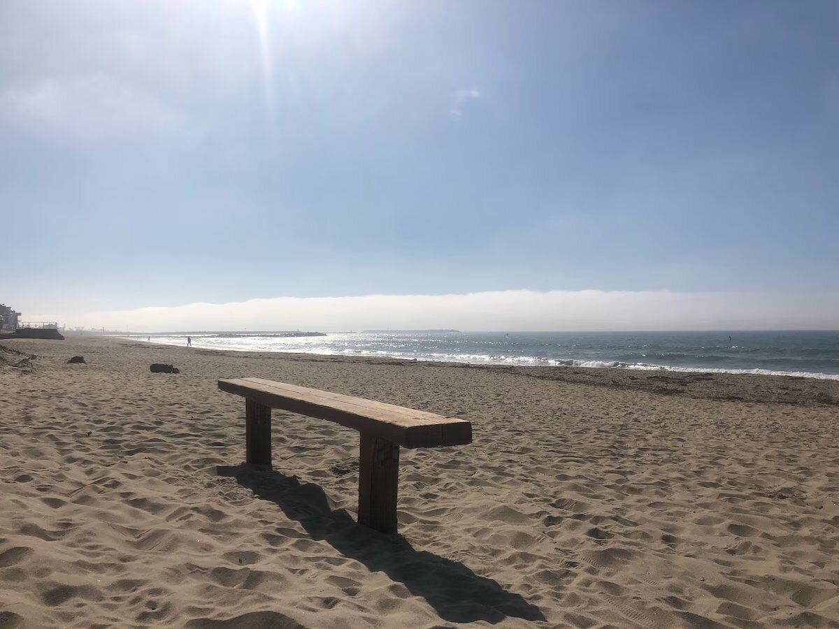 Ein sandiger Strand mit einer Holzbank und dem Meer im Hintergrund.