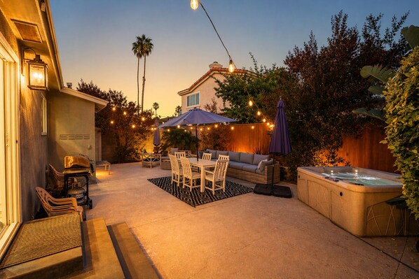 Un patio exterior con una mesa de comedor, sillas, un sofá, un jacuzzi, luces colgantes y una valla de madera.