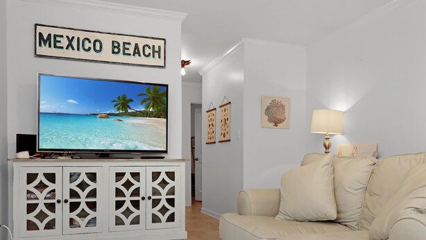 A living room with a television displaying a beach scene, a white cabinet with decorative cutouts, a beige sofa, and a sign reading "MEXICO BEACH". - 1st Street Bungalow (Port St. Joe)