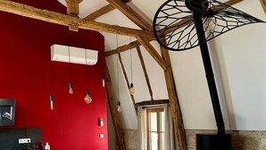A spacious loft with exposed wooden beams, a red accent wall, a kitchen area, a dining space, and a wood-burning stove. - Château de Belmont Ste Foi - a place steeped in history ! (Belmont-Sainte-Foi)