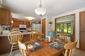 A dining area with a wooden table and chairs, a kitchen with wooden cabinets and appliances, and a view of greenery through large windows.