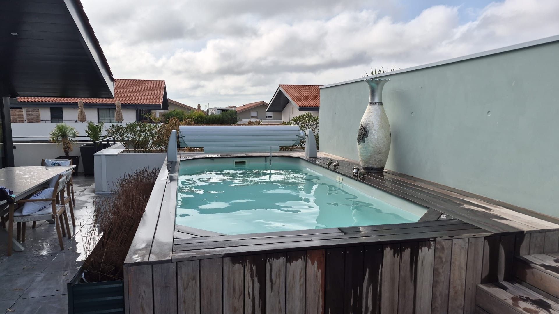 Ein Swimmingpool mit Holzterrasse, umgeben von einer niedrigen Mauer und einer großen Dekorationsvase.