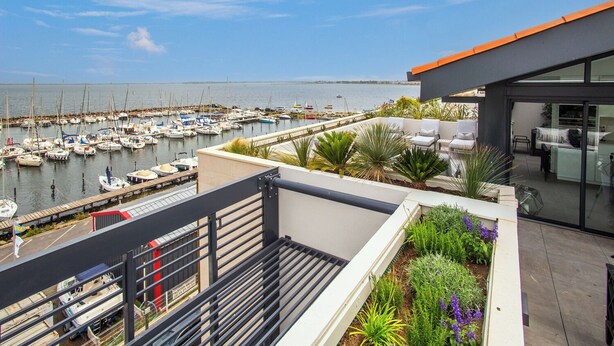 A rooftop terrace with a view of a marina, outdoor seating, and a modern building with large windows.