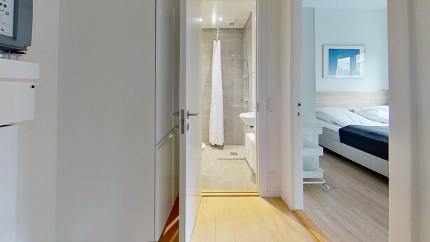 Una camera d'albergo moderna con pavimento in legno, un letto con biancheria bianca e blu, un bagno bianco con doccia e un piccolo lavandino.