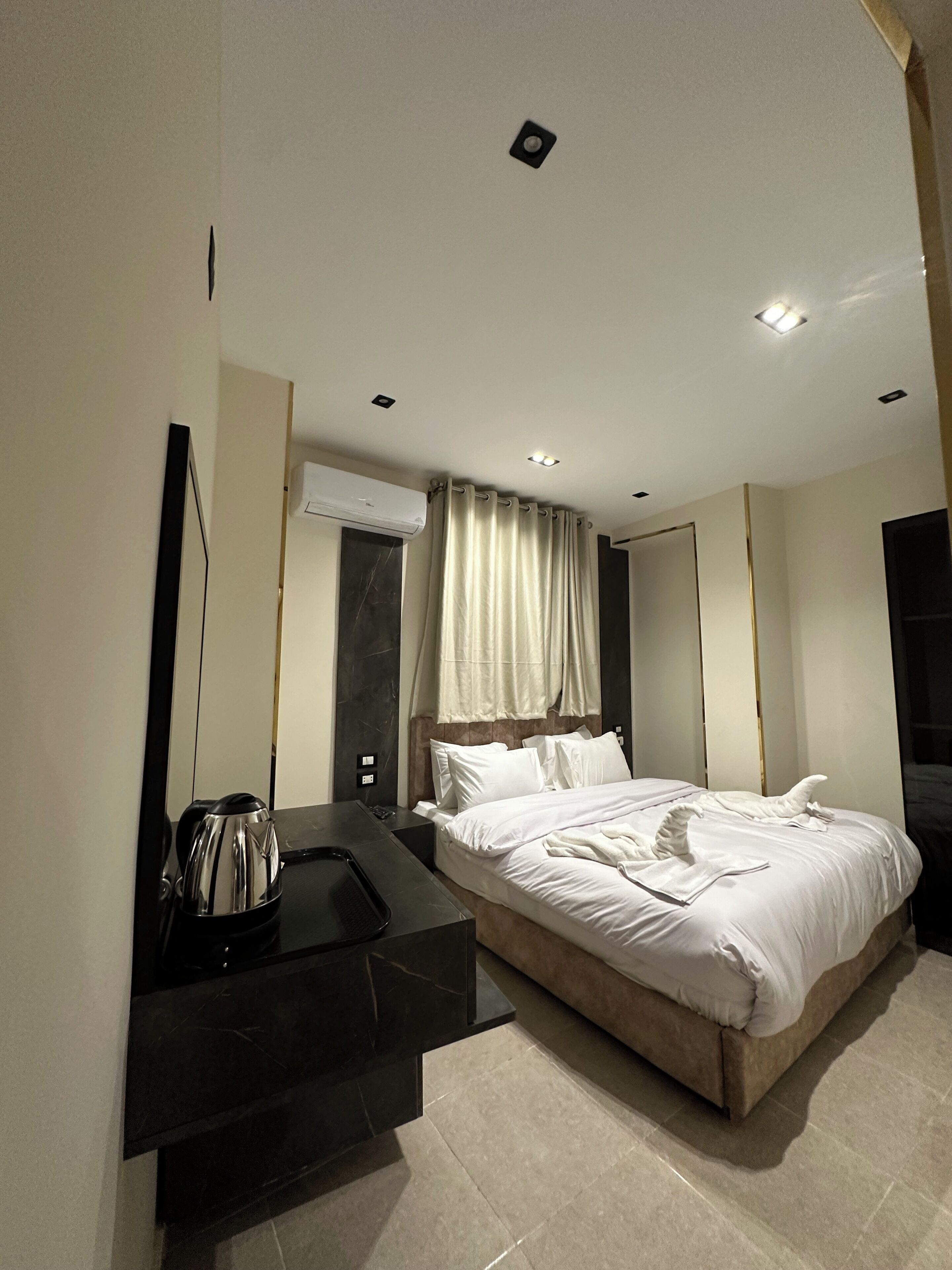 Une chambre d’hôtel moderne avec un grand lit, une table de chevet avec une bouilloire, un miroir et un luminaire au plafond.