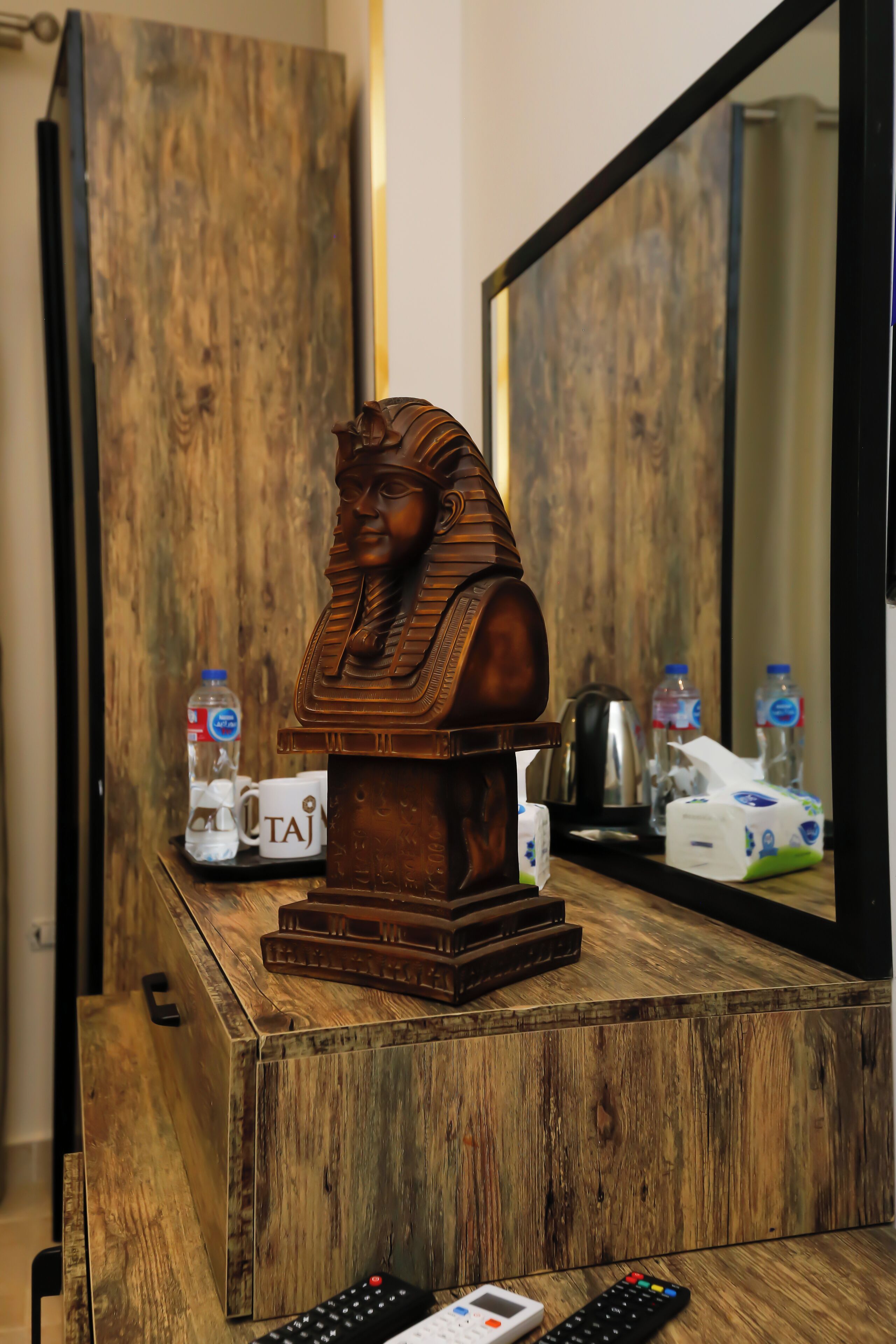 Un mobiletto in legno con un busto decorativo, un telecomando, una scatola di fazzoletti e una bottiglia d'acqua.