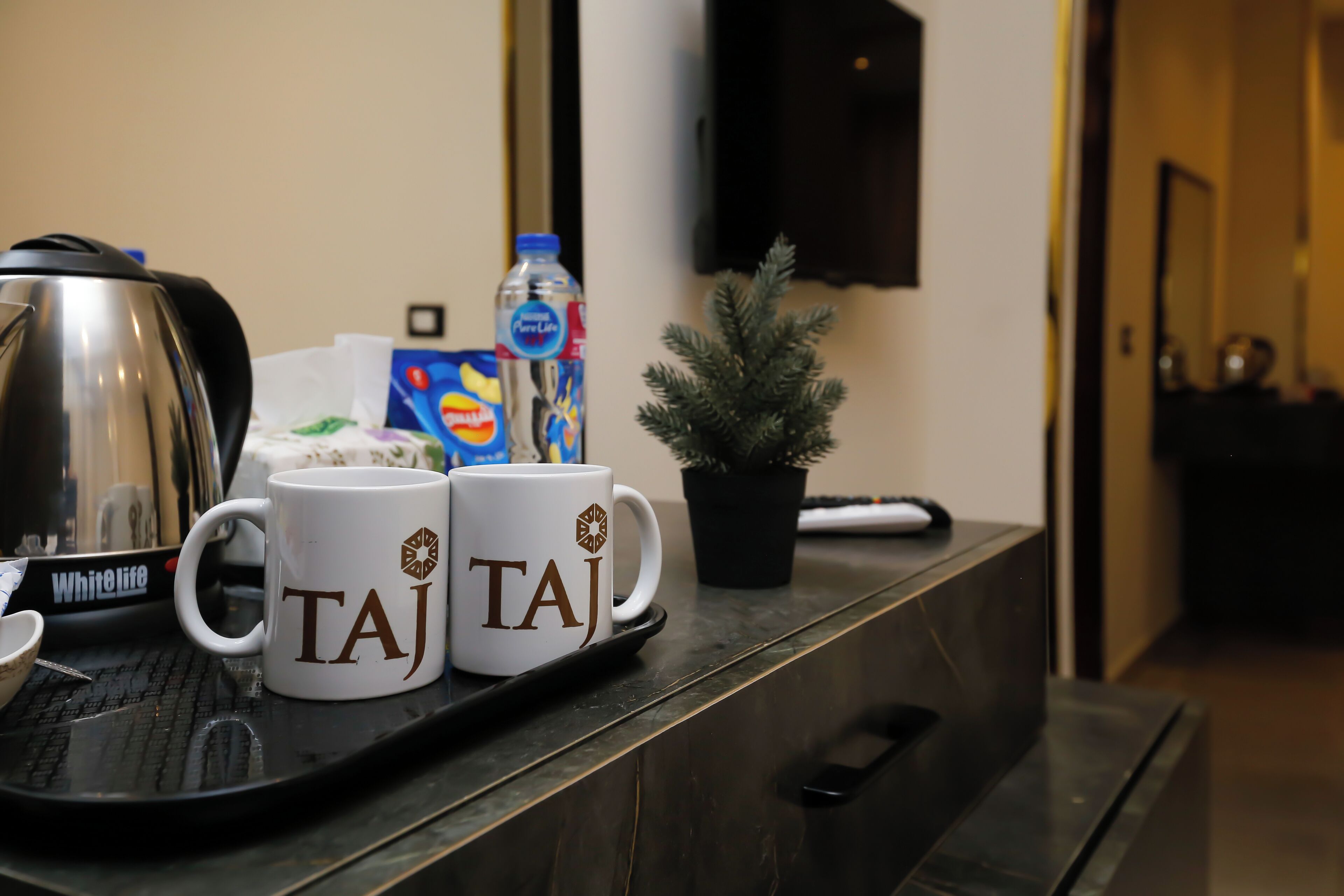 A tray with two white mugs labeled "TAJ" and a water bottle on a counter.