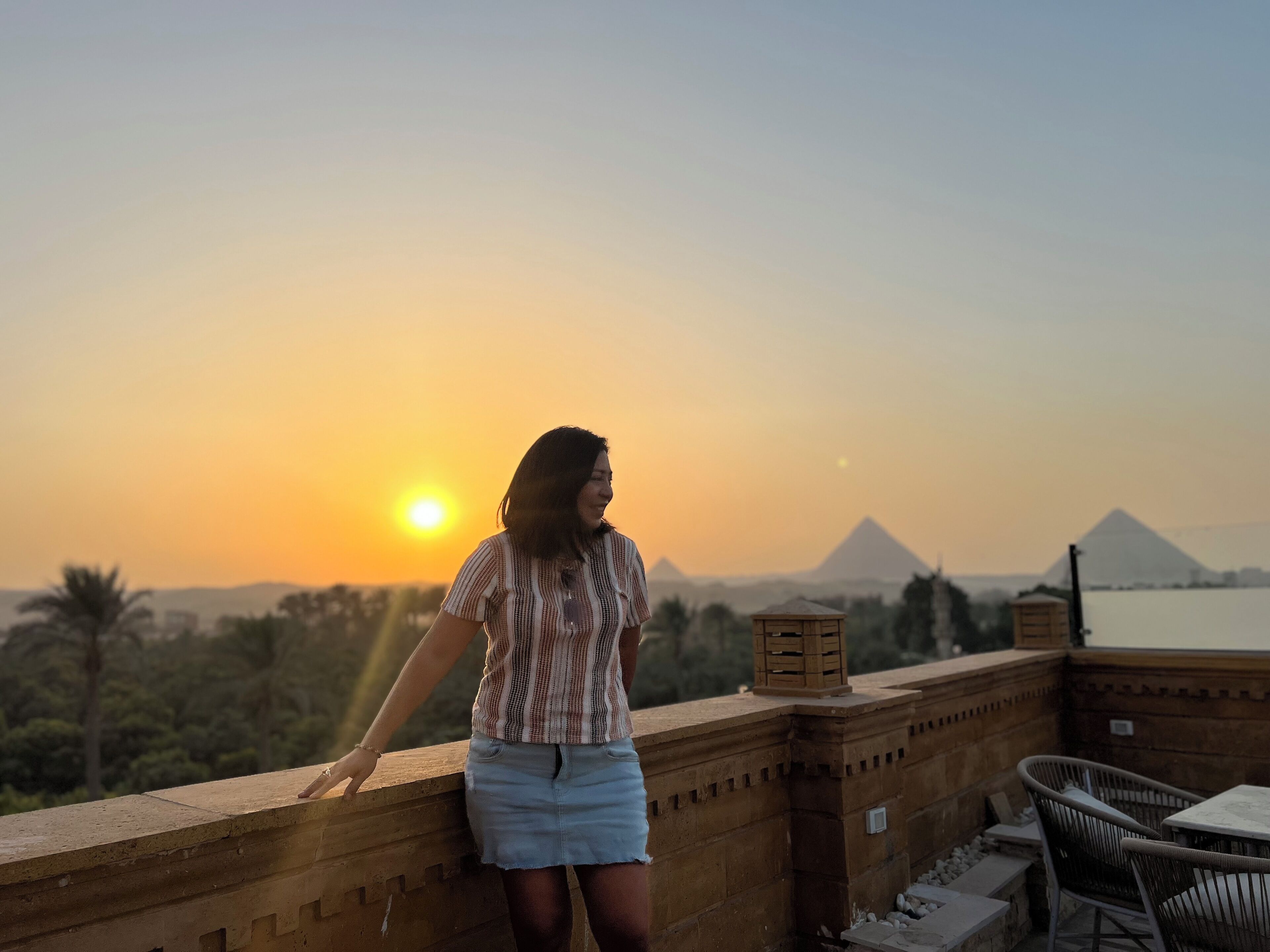 Eine Person steht auf einem Balkon und blickt auf einen Sonnenuntergang, im Hintergrund sind Pyramiden zu sehen.