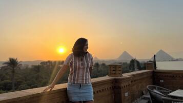 Eine Person steht auf einem Balkon und blickt auf einen Sonnenuntergang, im Hintergrund sind Pyramiden zu sehen.