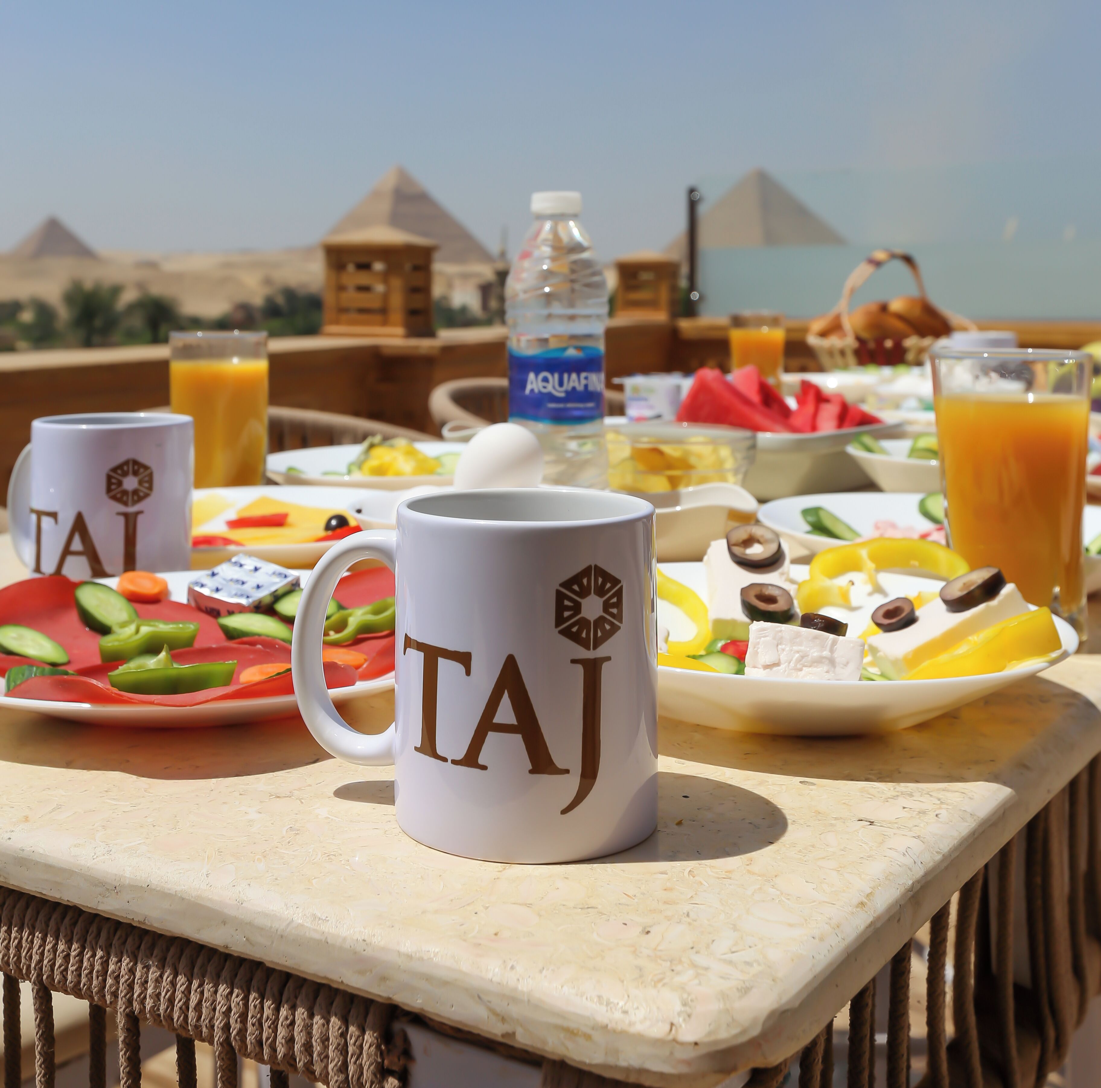 A breakfast table with a view of pyramids, featuring a Taj mug, plates of food, and a bottle of Aquafina water.