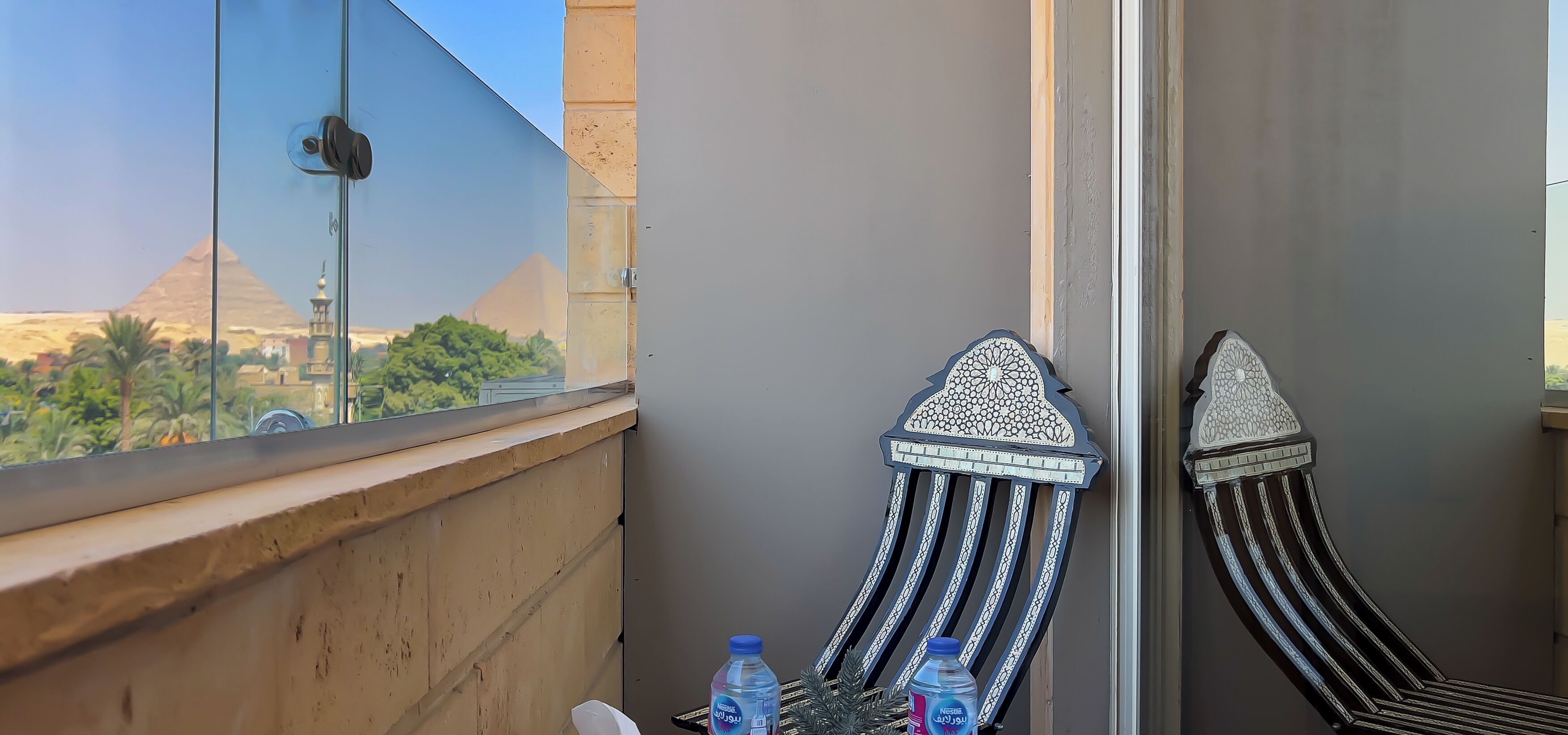 A balcony with a view of the pyramids, two intricately designed chairs, and a table with water bottles.