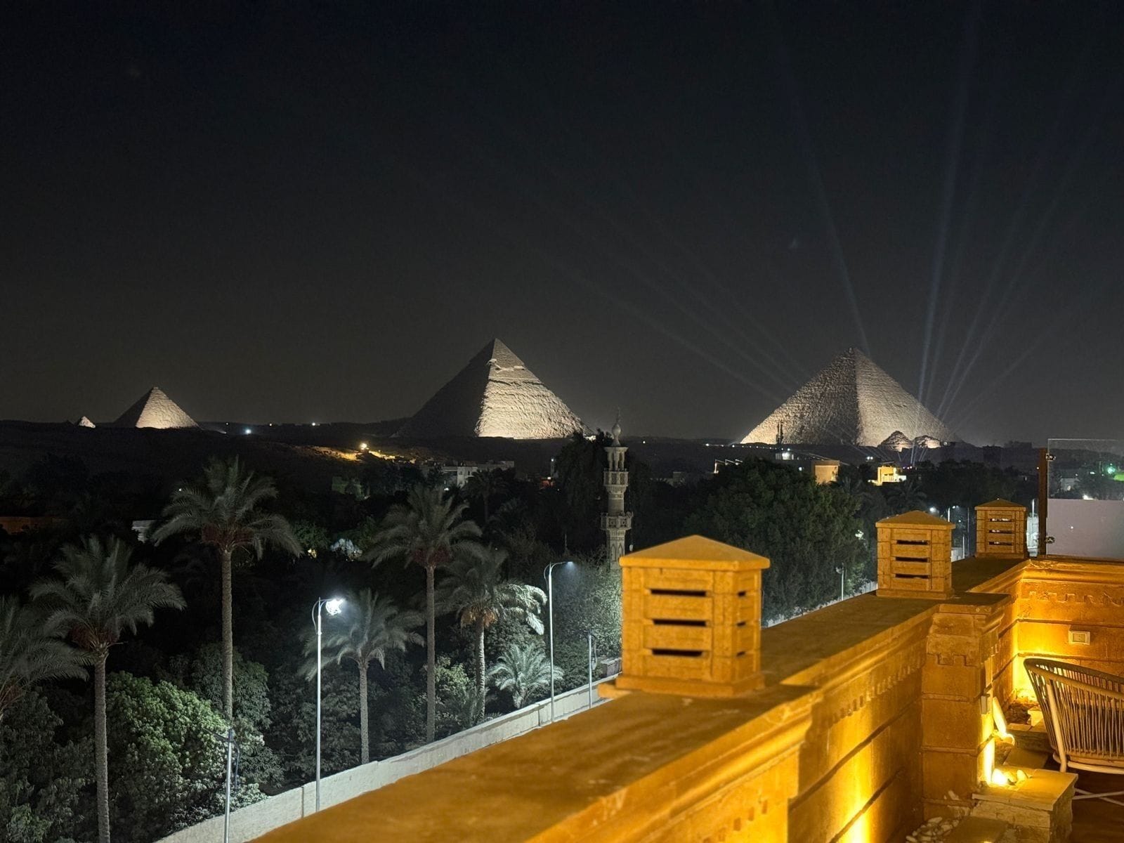 Una vista nocturna de una pirámide con una estructura iluminada y un camino bordeado de palmeras.