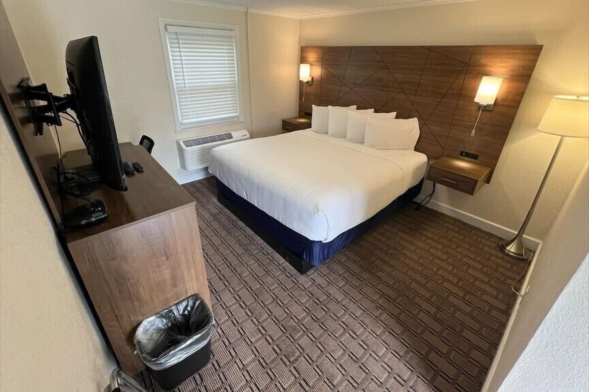 A hotel room with a bed, desk, computer, and lamp.