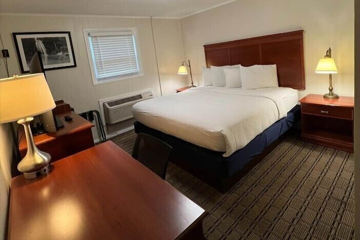 A hotel room with a bed, bedside tables, a desk, a chair, a lamp, a picture frame, a window with blinds, and a ceiling light.