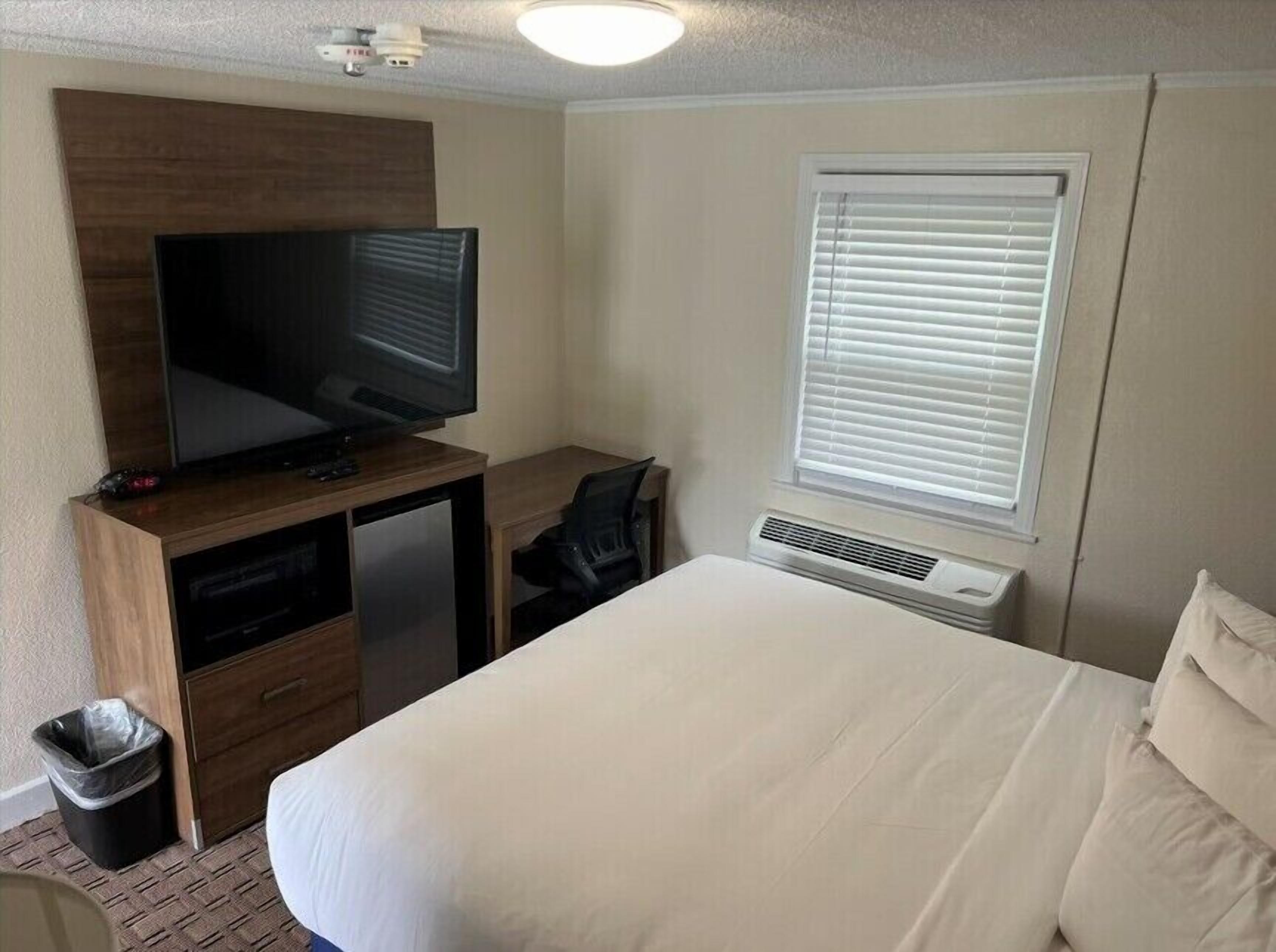 A hotel room with a bed, a desk with a television, a window with blinds, and a wall-mounted air conditioning unit.