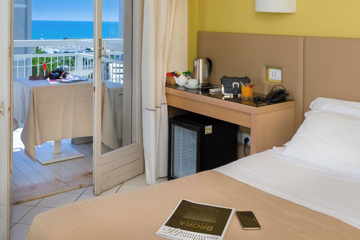 A hotel room with a bed, a desk with a kettle, a phone, and a small refrigerator.