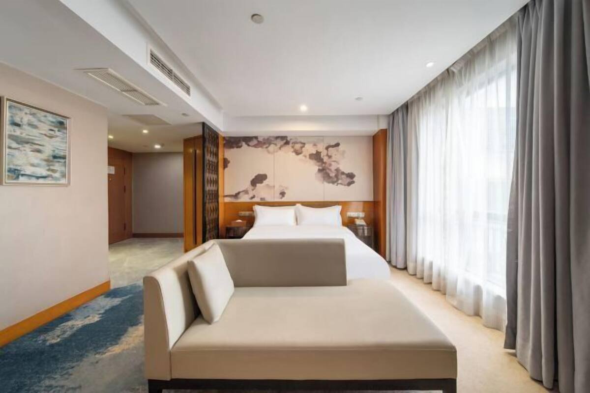 Een hotelkamer met een bed, een slaapbank, een groot raam met gordijnen, een schilderij aan de muur en een vloerbedekking.