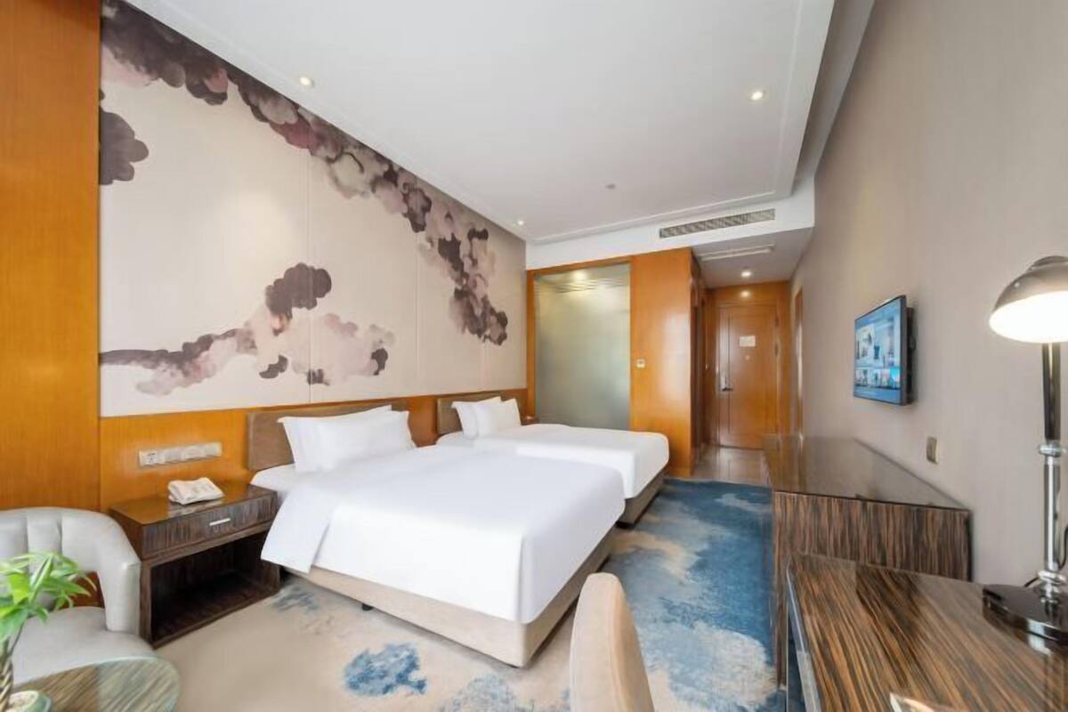 Een hotelkamer met twee bedden, een bureau, een stoel, een nachtkastje, een lamp en een muur met een wolkenfries.