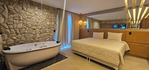 A modern bedroom with a stone wall, a large bed, a bathtub, and a mirror. - Pousada Alpes Azul (Campos do Jordão)