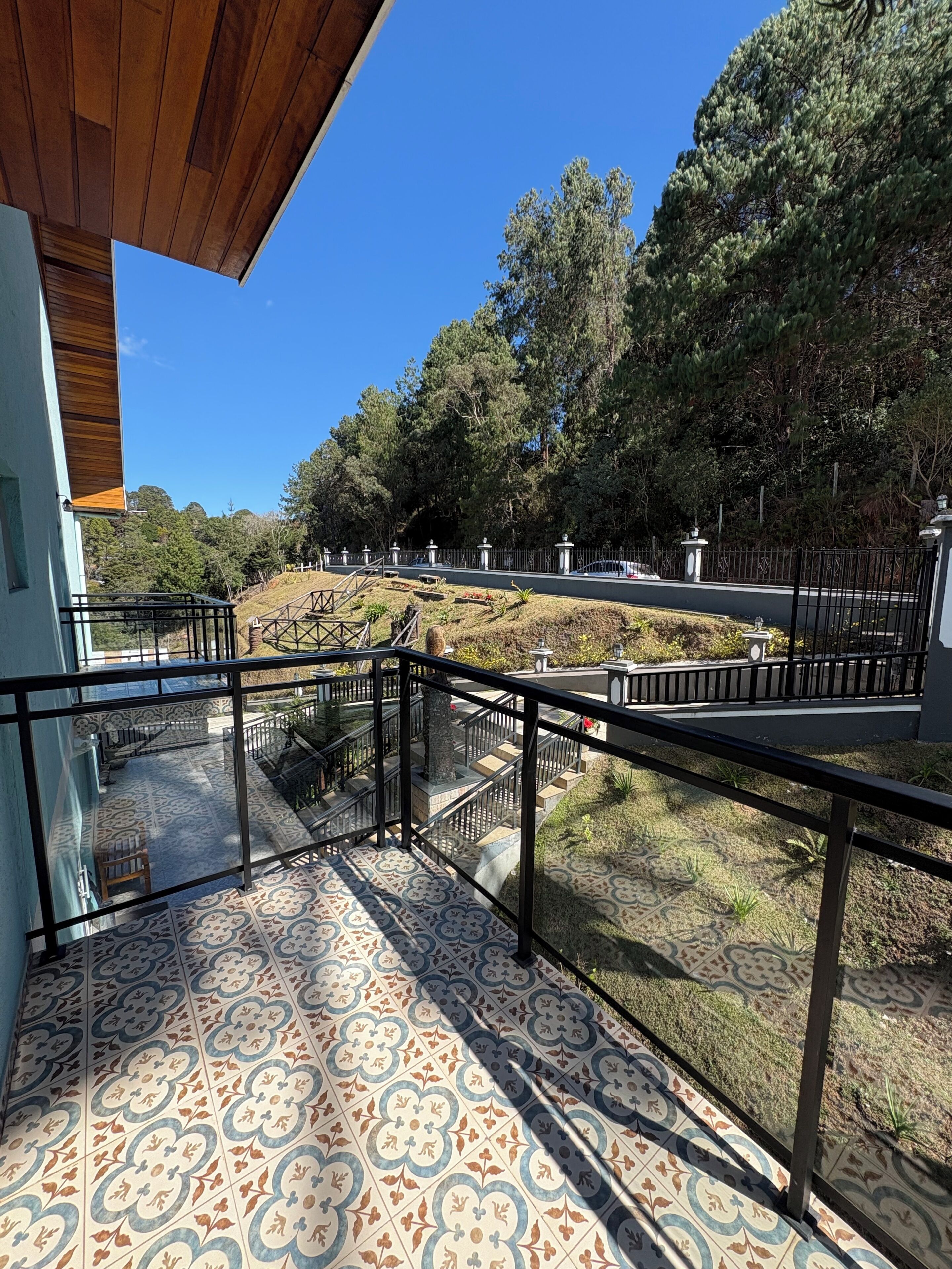 A balcony with patterned floor tiles, a railing, and a view of a landscaped area with trees and a fence.