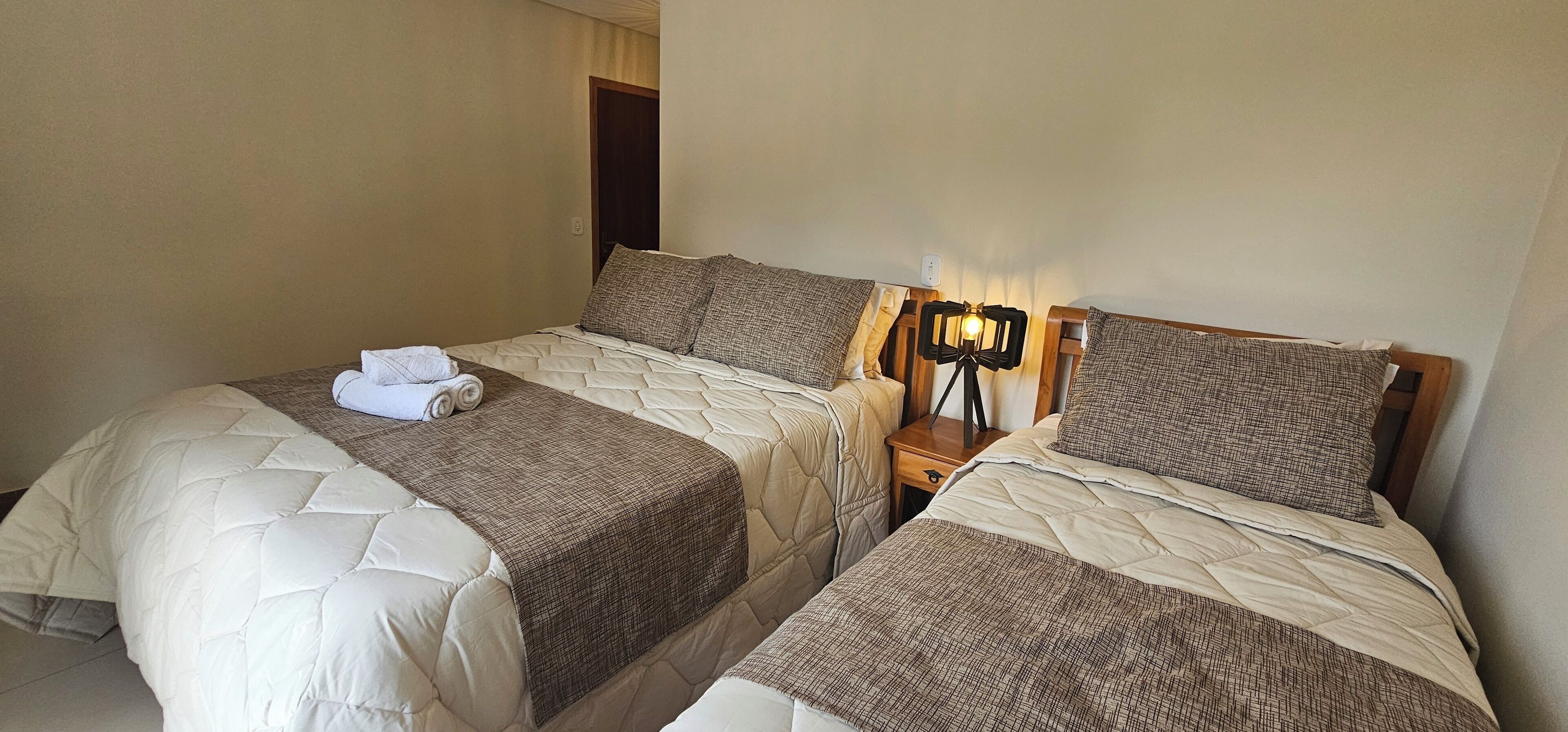 A hotel room with two beds, each with a quilted duvet and a brown checkered throw blanket. There is a bedside table with a lamp and a power outlet.