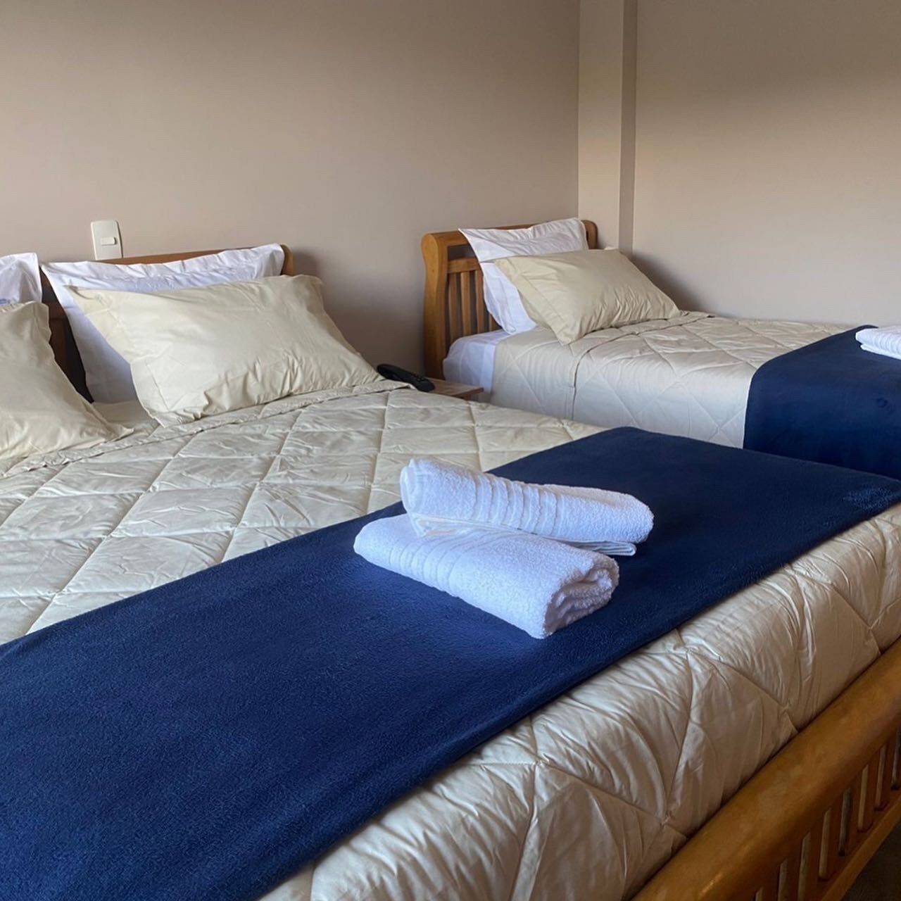 Habitación de hotel con dos camas, cada una con colchón acolchado y almohadas. Hay una manta azul en el suelo y una toalla doblada en la cama.
