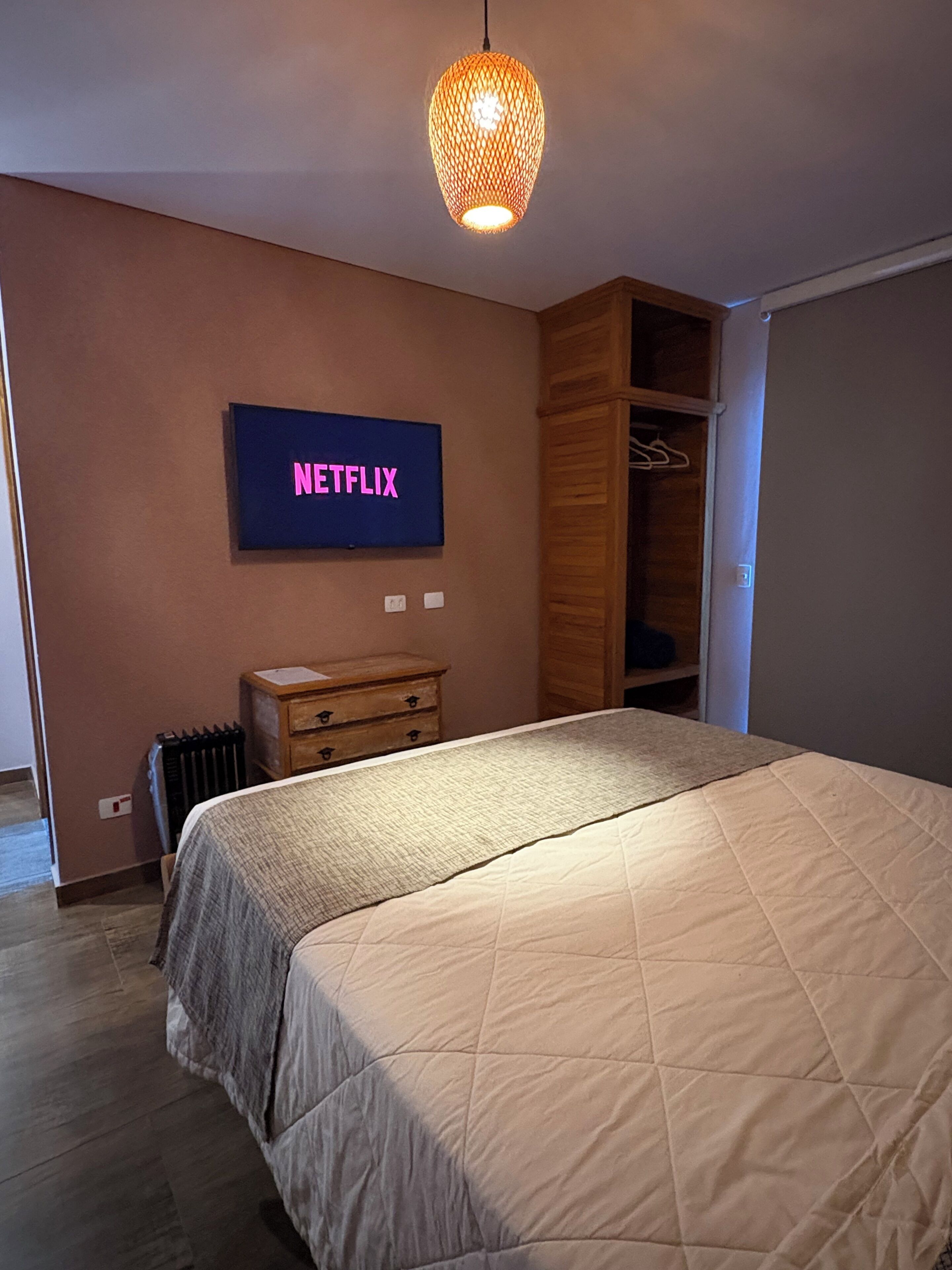 Un dormitori amb llit, armari, embutidora i radiador, i un televisor penjat a la paret que mostra 'NETFLIX'.