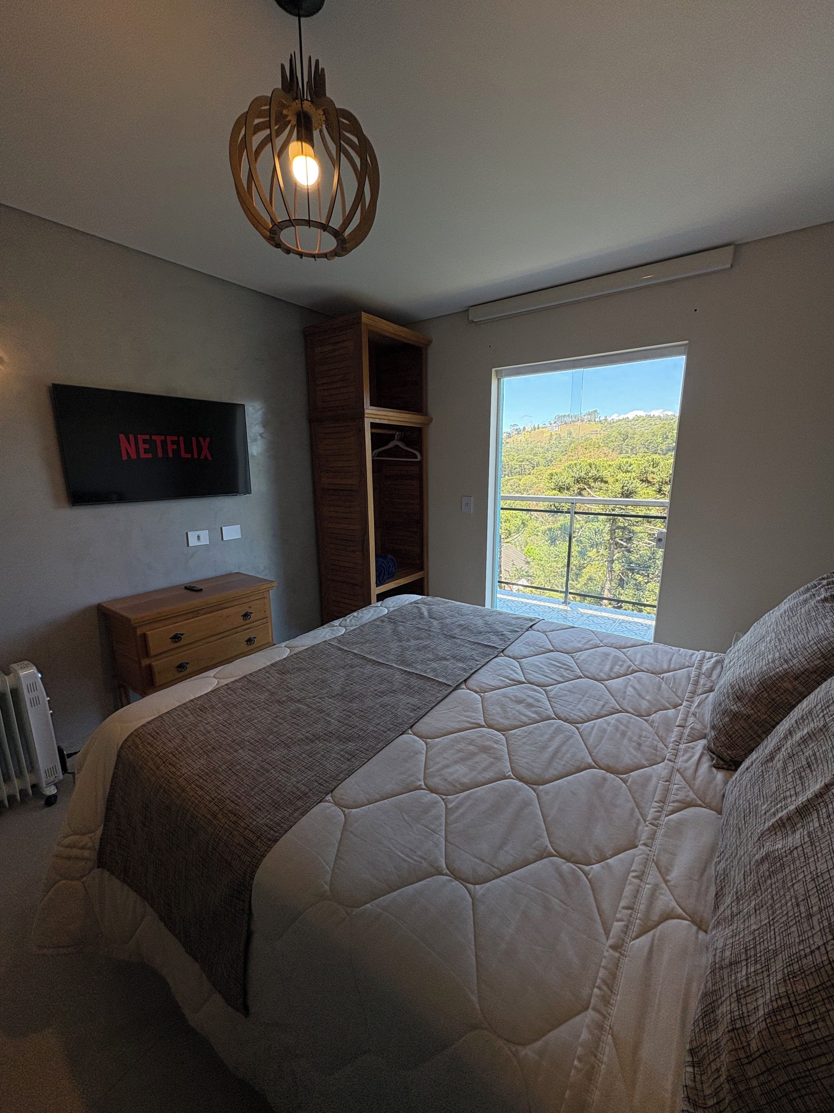 A bedroom with a bed, a wardrobe, a TV mounted on the wall, and a window with a view of greenery.