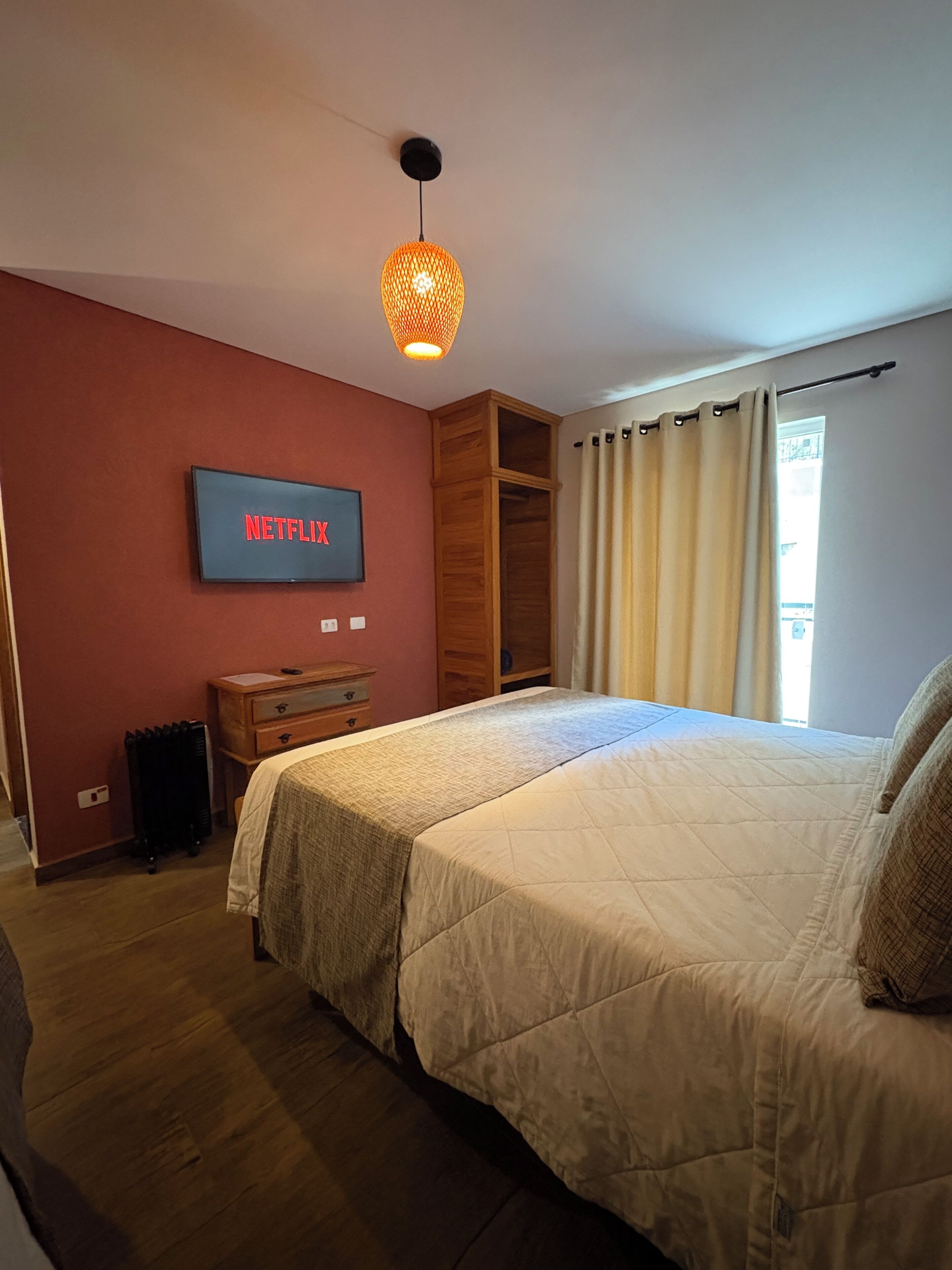 A bedroom with a bed, a TV mounted on the wall, a radiator, and a window with curtains.