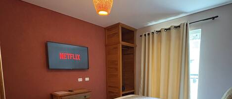 Un dormitorio con cama, televisor montado en la pared, radiador y ventana con cortinas.