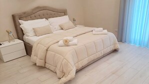 Un letto rifatto con un piumone beige e cuscini, un comodino con una lampada e vista su una tenda blu.