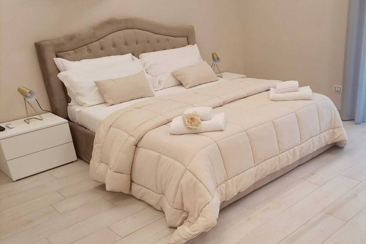 Cama bem arrumada com edredão e almofadas bege, mesa de cabeceira com abajur e vista para uma cortina azul.