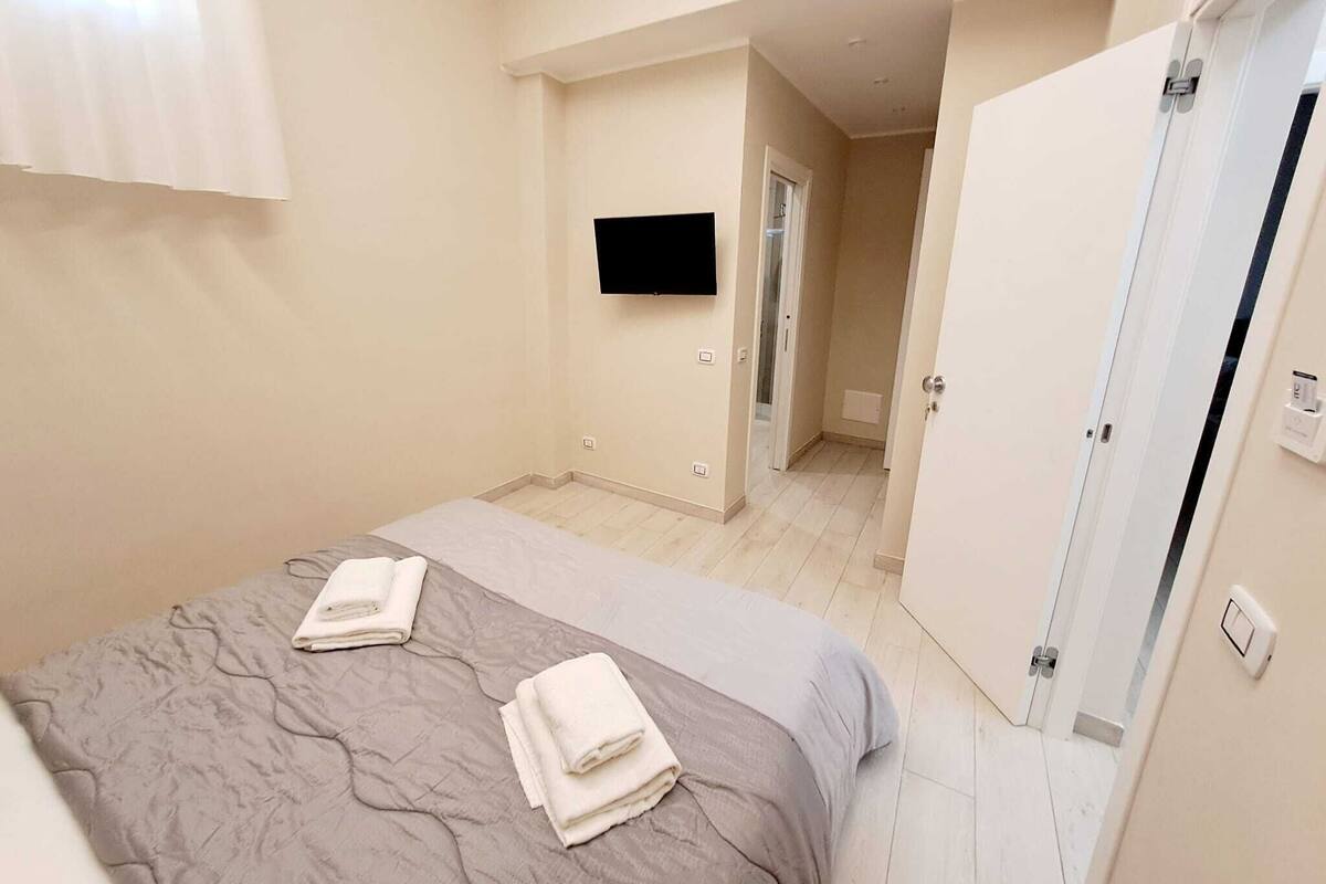 Quarto com uma cama, uma televisão presa na parede e uma porta que dá para outro cômodo.