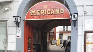 Ingresso a un edificio con arco rosso, con l'insegna "Mericano Pub & Bar" e l'indicazione dell'anno di fondazione: 2020.