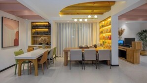 A modern dining area with a wooden ceiling, a white dining table, and chairs. There is a bookshelf with decorative items and a television on a wooden stand. - The Valley of Flowers (Lijiang)