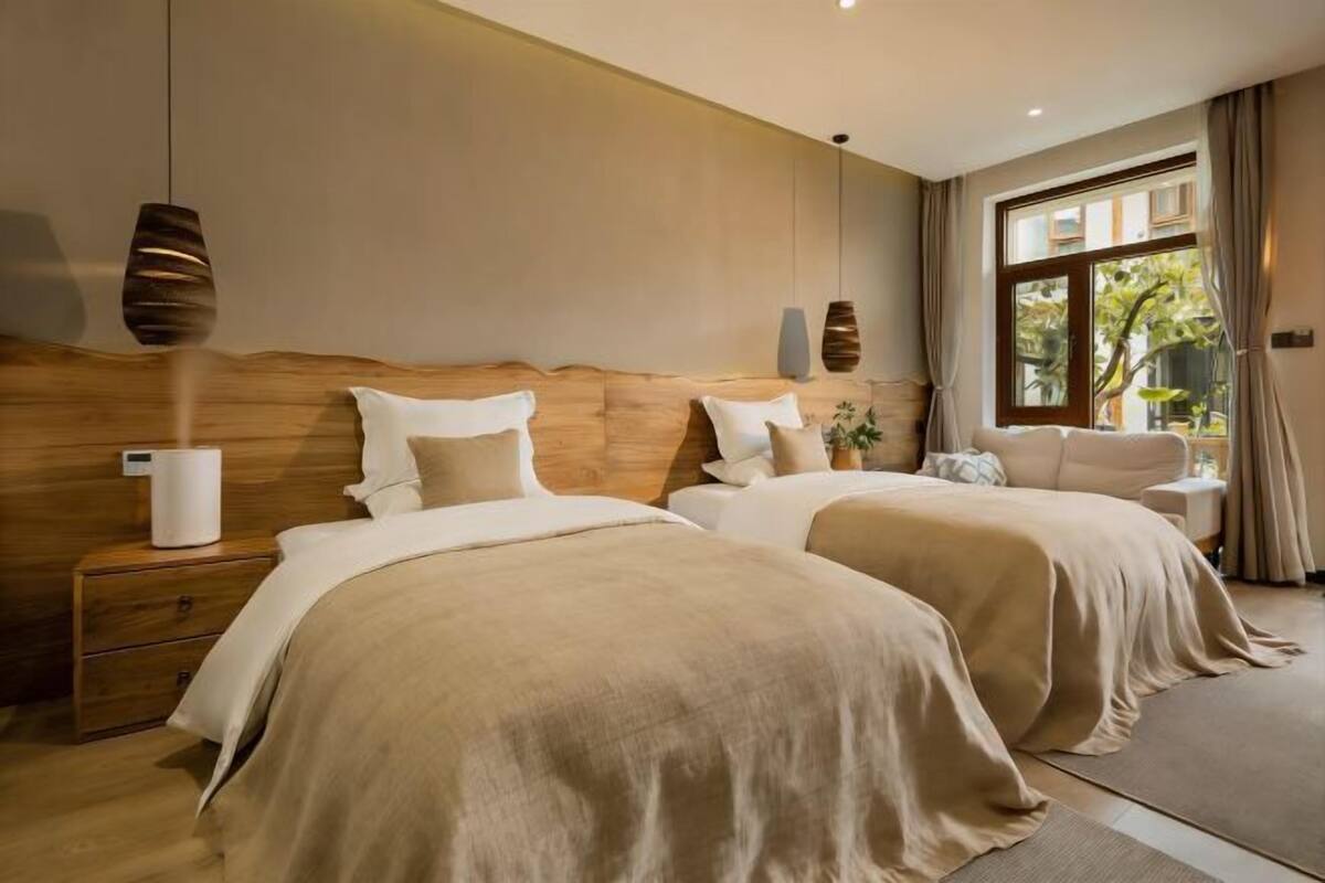 A hotel room with two beds, a wooden headboard, bedside tables, a nightstand, a window with curtains, and a view of greenery outside.