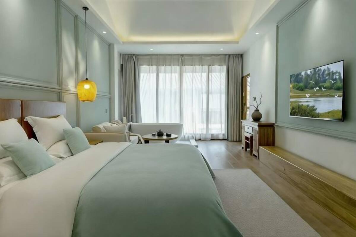 A hotel room with a large bed, a sitting area with a chair and table, a painting on the wall, and a view of a lake through the window.