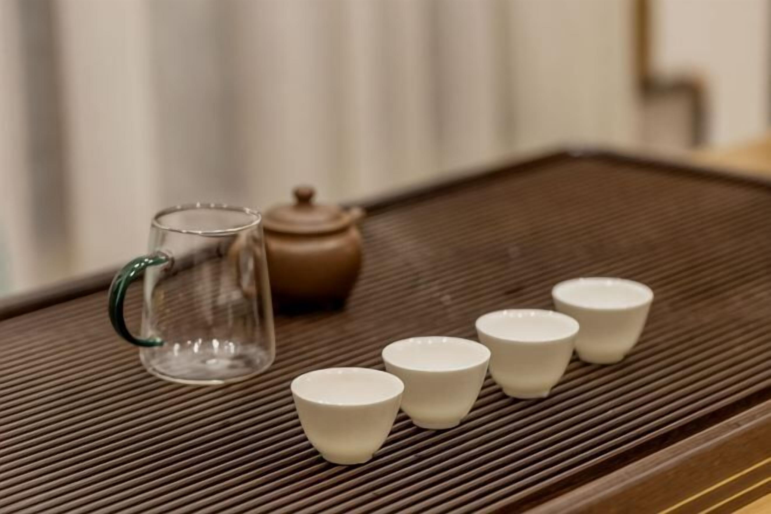 A clear glass pitcher, a brown teapot, and four white cups on a textured tray.