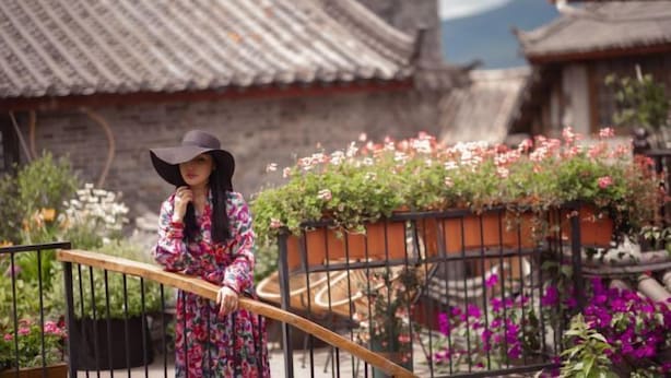 A person standing on a balcony with a traditional tiled roof and a railing. - The Valley of Flowers (Lijiang)
