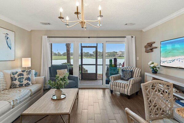 A spacious living room with a large window, a chandelier, and a television. - Driftwood Dreams BEACH condo (Miramar Beach)