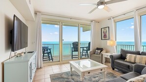 A spacious living room with a large TV, a ceiling fan, a sofa, a coffee table, and a view of the ocean through the sliding glass doors. - Direct Beachfront, Wrap Balcony, Breathtaking Views! Shores of Panama (Panama City Beach)
