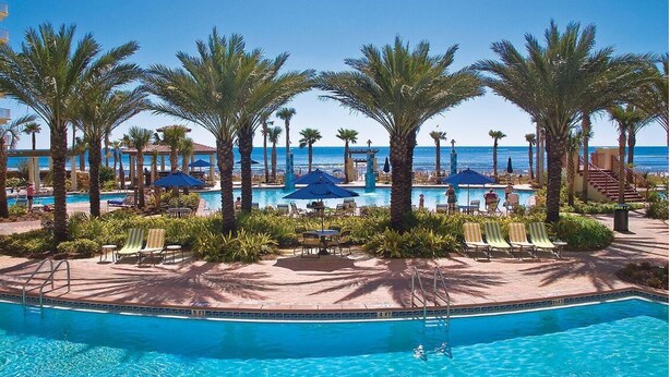 A resort pool area with palm trees, lounge chairs, and a view of the ocean. - Direct Beachfront, Wrap Balcony, Breathtaking Views! Shores of Panama (Panama City Beach)