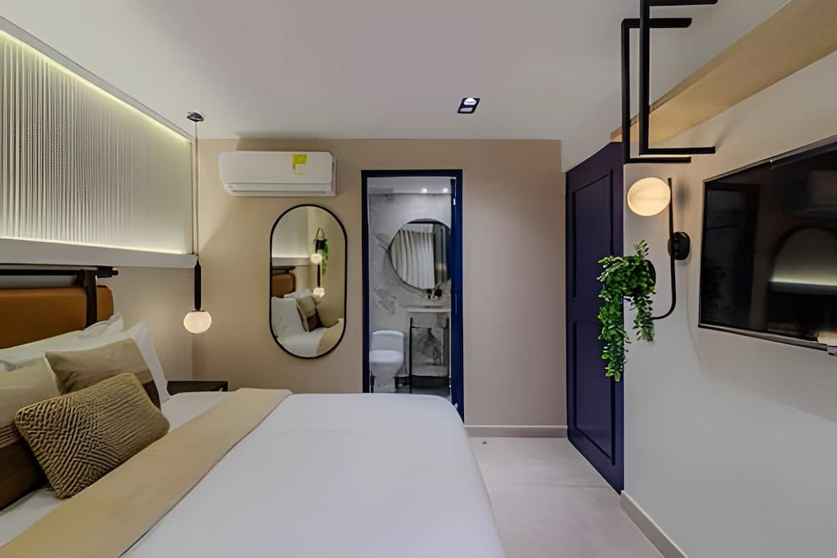 A modern bedroom with a bed, bedside tables, a mirror, an air conditioning unit, a wall-mounted TV, and a door leading to a bathroom.