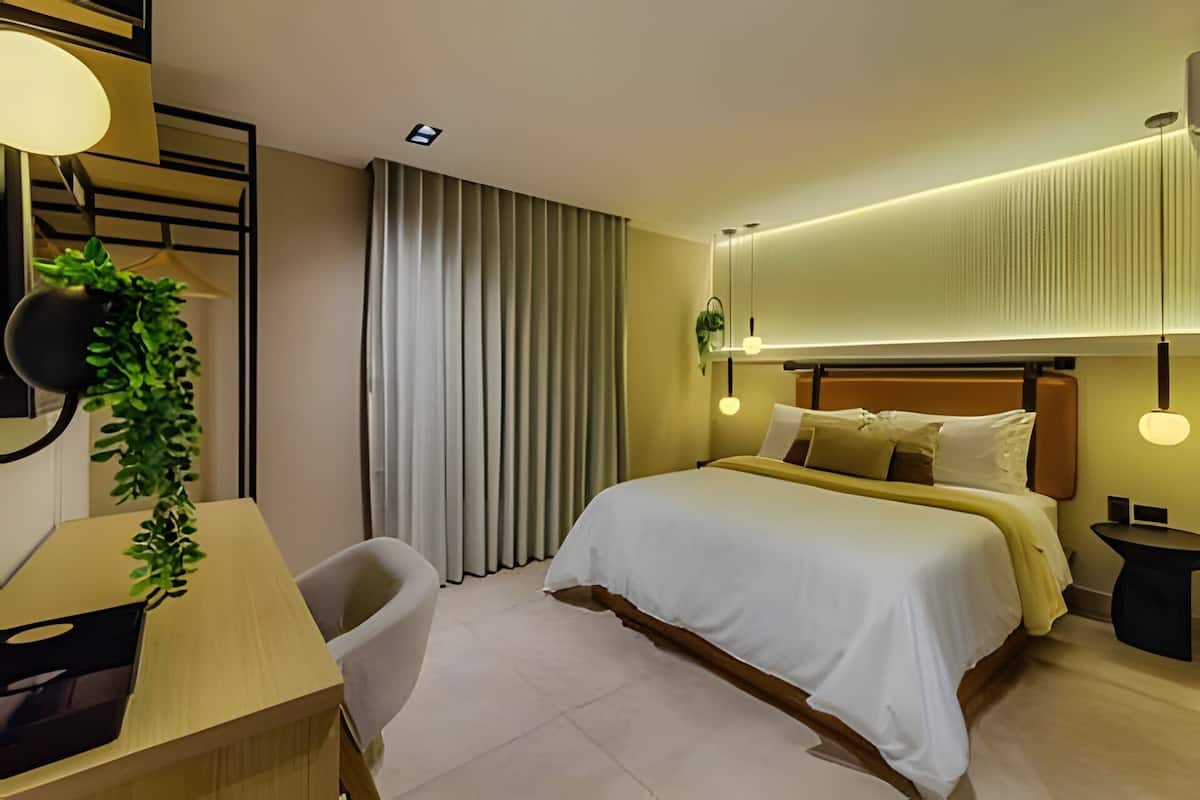 A modern hotel room with a bed, bedside table, chair, and a desk with a lamp.