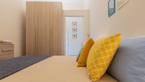 Un dormitorio con cama, dos almohadas, una mesita de noche, cómoda y una puerta que da a otra habitación.